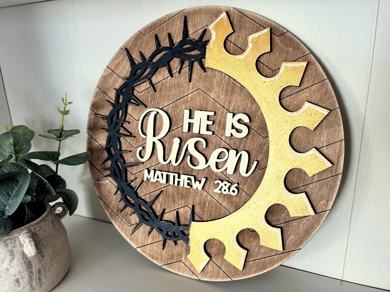 May include: A round wooden sign with a brown wood background and a chevron pattern. The sign features a black crown of thorns, a gold crown, and the words "He is Risen" with the scripture reference "Matthew 28:6".