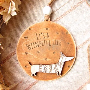May include: Wooden Christmas ornament with a dachshund design. The ornament is round with the text "It's a Wienerful Life" and a dachshund wearing a sweater and hat. The ornament is 3 inches in diameter.