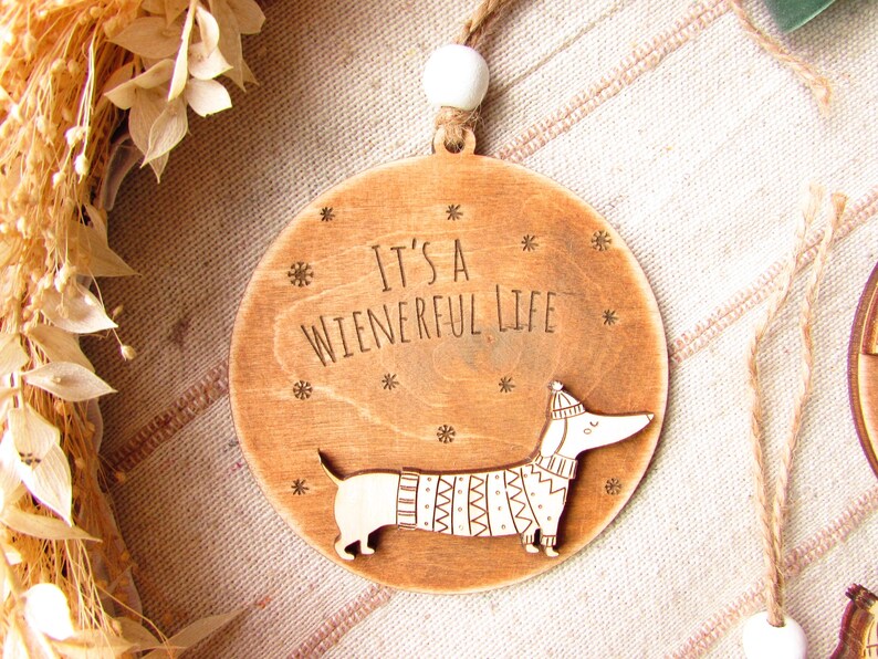 May include: Wooden Christmas ornament with a dachshund design. The ornament is round with the text "It's a Wienerful Life" and a dachshund wearing a sweater and hat. The ornament is 3 inches in diameter.