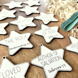 May include: A collection of white wooden star ornaments with black lettering. Each star features a different Bible verse, including "Jesus", "The Rock", "Author of Salvation", "Lamb of God", and "Prince of Peace".