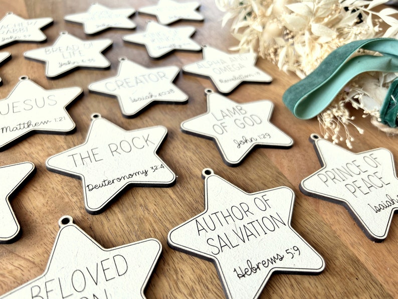 May include: A collection of white wooden star ornaments with black lettering. Each star features a different Bible verse, including "Jesus", "The Rock", "Author of Salvation", "Lamb of God", and "Prince of Peace".