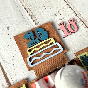May include: A wooden birthday cake with the number 10 on top. The cake is decorated with blue, green, and yellow frosting. There is a separate wooden number 10 with a crown on top.