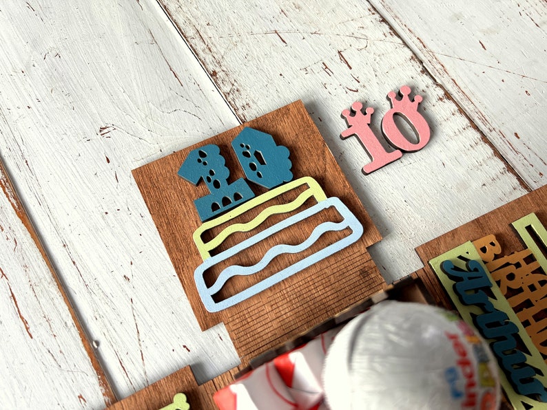 May include: A wooden birthday cake with the number 10 on top. The cake is decorated with blue, green, and yellow frosting. There is a separate wooden number 10 with a crown on top.