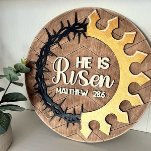 May include: A round wooden sign with a brown wood grain background. The sign features a black crown of thorns, a gold crown, and the words "He is Risen" with the scripture reference "Matthew 28:6".