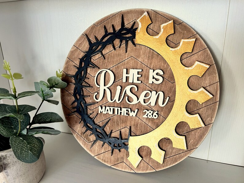 May include: A round wooden sign with a brown wood grain background. The sign features a black crown of thorns, a gold crown, and the words "He is Risen" with the scripture reference "Matthew 28:6".