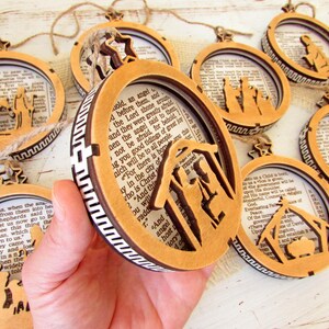 May include: Wooden Christmas ornaments with biblical scenes. Each ornament features a laser-cut design of a nativity scene or related figures, set against a backdrop of printed text. The ornaments are circular or oval-shaped with a star-shaped top.
