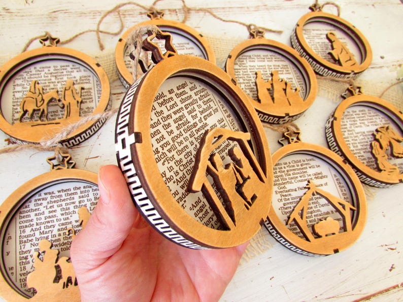 May include: Wooden Christmas ornaments with biblical scenes. Each ornament features a laser-cut design of a nativity scene or related figures, set against a backdrop of printed text. The ornaments are circular or oval-shaped with a star-shaped top.