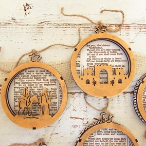 May include: Wooden Christmas ornaments with a cutout design featuring biblical scenes. Each ornament has a circular shape with a star-shaped top and is strung with twine. The background of each ornament is a page from a book.