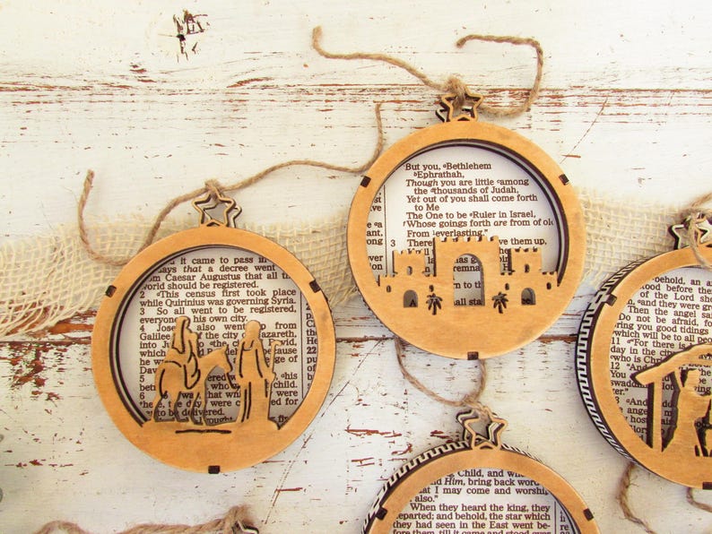 May include: Wooden Christmas ornaments with a cutout design featuring biblical scenes. Each ornament has a circular shape with a star-shaped top and is strung with twine. The background of each ornament is a page from a book.