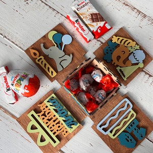 May include: A wooden box with a birthday theme. The box is decorated with colorful illustrations of an elephant, balloons, a cake, and a party hat. The box is open and filled with chocolate candies wrapped in red foil.