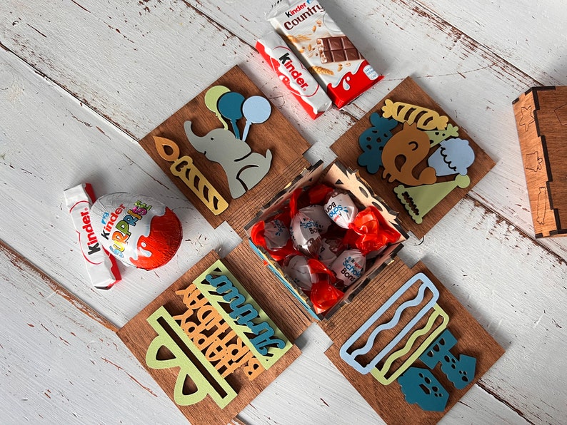 May include: A wooden box with a birthday theme. The box is decorated with colorful illustrations of an elephant, balloons, a cake, and a party hat. The box is open and filled with chocolate candies wrapped in red foil.