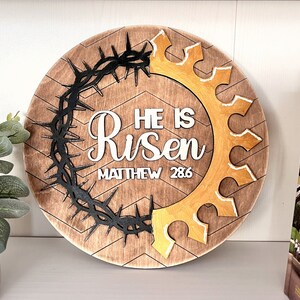 May include: A round wooden sign with a chevron pattern. The sign features a black crown of thorns, a gold crown, and the words "He is Risen" with the scripture reference "Matthew 28:6".