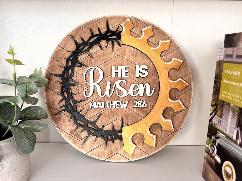 May include: A round wooden sign with a chevron pattern. The sign features a black crown of thorns, a gold crown, and the words "He is Risen" with the scripture reference "Matthew 28:6".