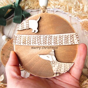 May include: Wooden Christmas ornament with a dachshund dog design. The ornament is round and features the text "merry christmas". The dog is wearing a sweater and hat. The ornament is held in a hand.