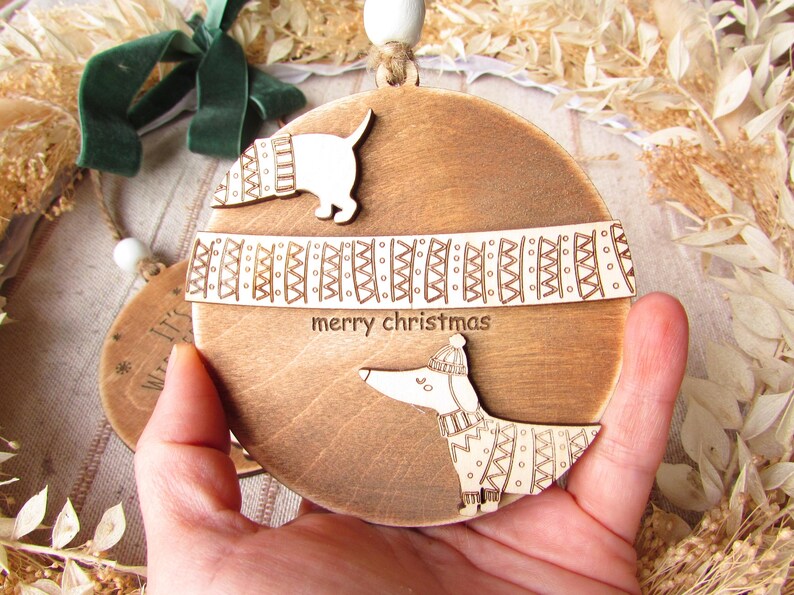 May include: Wooden Christmas ornament with a dachshund dog design. The ornament is round and features the text "merry christmas". The dog is wearing a sweater and hat. The ornament is held in a hand.