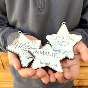 May include: Three white wooden star ornaments with black lettering. The ornaments read "Prince of Peace" with Isaiah 9:6, "Immanuel" with Isaiah 7:14, and "Alpha and Omega" with Revelation 1:8.