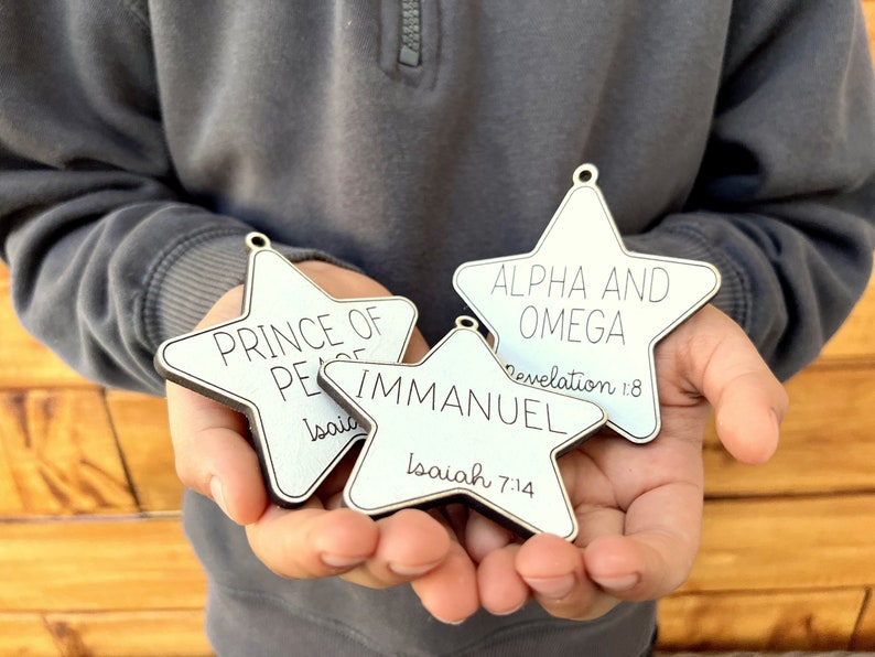 May include: Three white wooden star ornaments with black lettering. The ornaments read "Prince of Peace" with Isaiah 9:6, "Immanuel" with Isaiah 7:14, and "Alpha and Omega" with Revelation 1:8.