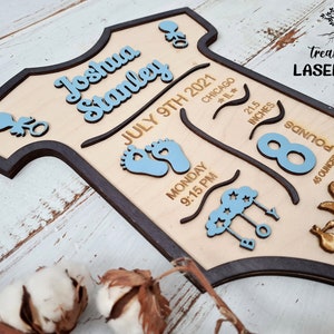 May include: A wooden baby onesie-shaped sign with the name Joshua Stanley, birthdate July 9th, 2021, time 9:15 PM, location Chicago, IL, weight 8 pounds, 45 ounces, and length 21.5 inches. The sign is painted in blue and brown.