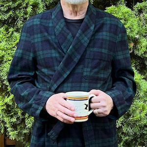 May include: A long-sleeved robe featuring a dark green and navy blue plaid pattern with a shawl collar, a tie waist, and two front pockets. The robe is worn over a black shirt and blue jeans, and a white ceramic mug with a bird graphic is held.