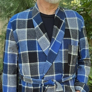 May include: A long-length robe featuring a blue, black, and grey plaid pattern. The garment includes a shawl collar, long sleeves, two front patch pockets, and a matching fabric tie belt secured at the waist over a black shirt and blue jeans.