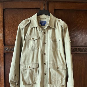 May include: Tan-colored jacket with multiple pockets and button closures. The jacket has a collared neckline and epaulettes on the shoulders. The brand name "Pendleton" is visible on a label. The jacket is hanging on a black hanger against a wooden background.