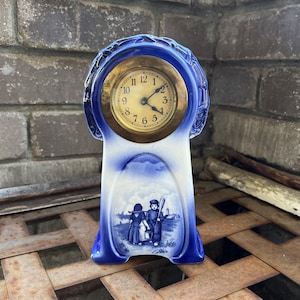 May include: A vintage blue and white ceramic mantel clock with a gold-toned clock face. The clock features a decorative design with a pastoral scene. The clock measures approximately 8 inches tall.