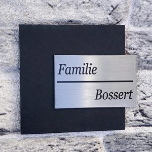 Könnte beinhalten: Eine quadratische schwarze Schieferplatte mit einer rechteckigen, gebürsteten silbernen Metallplatte auf der Vorderseite. Die Metallplatte zeigt den Text Familie in der oberen Zeile und Bossert in der unteren Zeile, getrennt durch eine schwarze Linie.