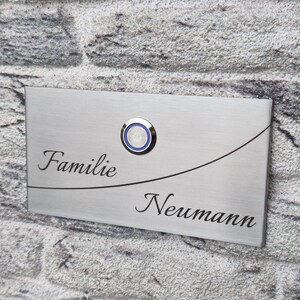 Könnte beinhalten: Eine rechteckige Türklingelplatte aus gebürstetem Edelstahl mit einem runden Druckknopf und blauem LED-Ring. Die Platte ist mit dem Text "Familie" oben links und "Neumann" unten rechts graviert, getrennt durch eine geschwungene Linie.