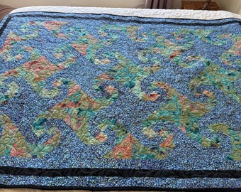 May include: A large, quilted bedspread with a blue floral pattern and a black border. The quilt features a repeating design in shades of blue, green, and peach. The bedspread is made of a soft, quilted material.