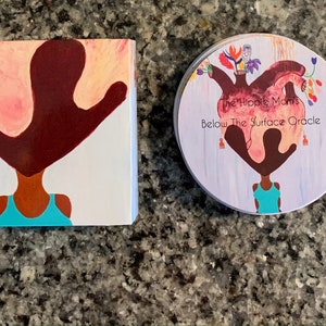 May include: A round oracle deck box with a purple, pink, and white background. The box features a painting of a woman with a heart on her head. The text "The Hippie Moms Below The Surface Oracle" is printed on the box.