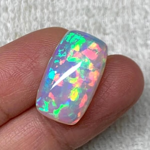 Puede incluir: Una piedra preciosa de ópalo rectangular con una superficie pulida. El ópalo muestra un vibrante juego de colores, con destellos de verde, rosa, amarillo y azul. La piedra se sostiene entre los dedos, mostrando su tamaño y forma. La gema es un hermoso ejemplo de ópalo natural.