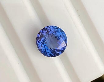 May include: A round, faceted gemstone displaying a vibrant gradient of deep blue and violet hues. The stone features a precise, symmetrical cut with numerous triangular facets that reflect light, creating a brilliant, sparkling appearance.