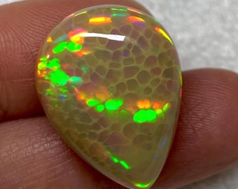 May include: A pear-shaped opal gemstone with a unique honeycomb pattern. The stone displays a range of colors, including green, yellow, orange, and purple, with a polished, glossy surface. The opal is held by a finger.