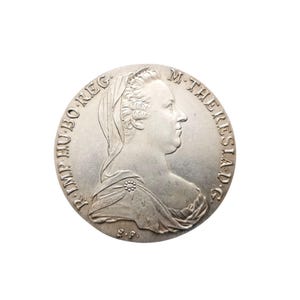 May include: A silver coin featuring a profile of a woman wearing a veil and adorned with a floral brooch. The coin has inscriptions around the edge, and the overall design is detailed and historical.
