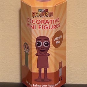 May include: A hexagonal box featuring the text "ITALIAN BRAINROT DECORATIVE MINI FIGURES" and cartoon figures. The box is predominantly orange and brown, with the words "Attach Me!" displayed on the side.