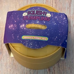 May include: A round, tan-coloured sensory fidget toy shaped like a dumpling steamer, enclosed in a purple paper sleeve with a starry night pattern. The sleeve features the text "SQUISHY DUMPLING" and "sensory fidget toy" in white and yellow.