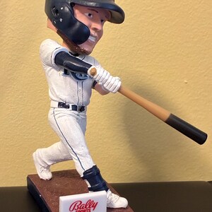 May include: A bobblehead figurine of a baseball player in a white uniform with blue accents, wearing a navy blue helmet. The figurine is of Austin Meadows, with the Bally Sports Sun logo on the base.