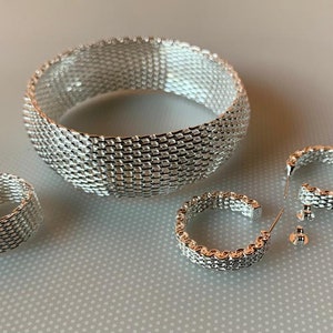 May include: A set of silver jewellery featuring a wide bangle bracelet, a ring, and a pair of hoop earrings. All pieces are made of a delicate, interwoven chain design.