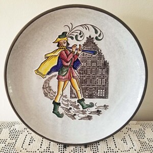 May include: A round, white ceramic plate with a brown rim, decorated with a colourful illustration. The design shows a figure in medieval clothing playing a flute, followed by mice, with a building in the background.