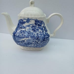 May include: A white ceramic teapot featuring a blue transferware pattern depicting a rural landscape with a person, trees, and a building. The teapot has a curved handle, a spout, and a matching lid with a small knob handle on top.