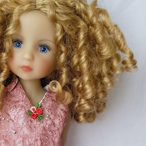 Peut inclure: Une poupée aux longs cheveux blonds bouclés portant une robe rose avec des détails en dentelle et un nœud blanc. La poupée a les yeux bleus et porte des chaussures roses.