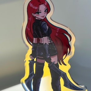 May include: An acrylic standee featuring an anime-style character with long red hair, wearing a black cropped top, shorts, and dark armour pieces on the shoulders and forearms. The character holds two curved blades and has a tattoo on the side.