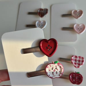 Op de afbeelding: Een collectie haarspeldjes met knopen in de vorm van hartjes en appeltjes. De speldjes zijn bevestigd op witte presentatiekaartjes. De ontwerpen omvatten een effen rood hart, een witte appel met rode bloemmotieven en diverse geruite hartjes.