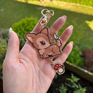 Op de afbeelding: Een heldere acryl sleutelhanger met een cartoon hertje, een vlinder en een rode paddenstoel bedel. Het hertje is bruin met een witte buik en zwarte ogen. De sleutelhanger heeft een goudkleurige hartvormige sluiting.