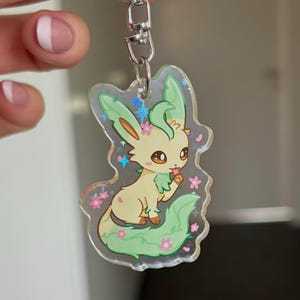 Op de afbeelding: Een heldere acryl sleutelhanger met een cartoonachtig wezen met groen en geel haar, groene oren en een bladachtige staart. Roze bloemen en blauwe sterren versieren de randen. De sleutelhanger is bevestigd aan een zilveren metalen sluiting.