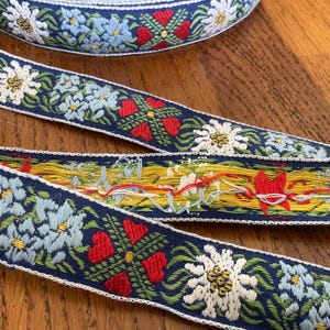 May include: Close-up of decorative embroidered ribbon with a navy blue background. The ribbon features a repeating pattern of white flowers, light blue flowers, and red hearts. A section of yellow and red thread is visible.