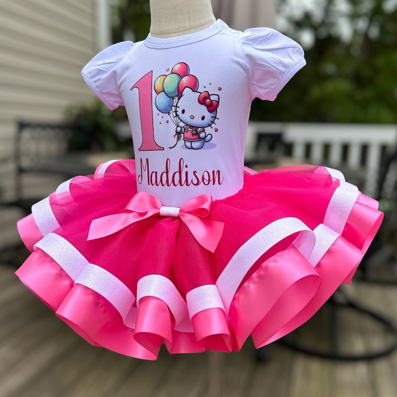 May include: A pink tulle tutu with white satin ribbon trim. A white t-shirt with a Hello Kitty graphic and the number 1, along with the name "Maddison" is worn over the tutu.