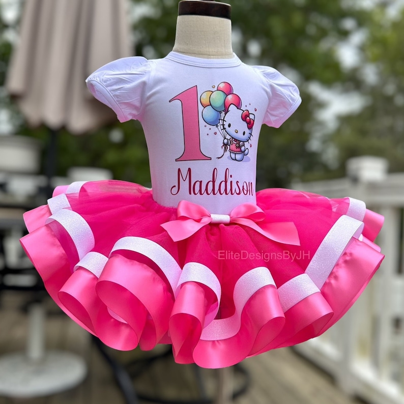 May include: A pink tulle tutu with white satin ribbon trim. A white t-shirt with a pink number one, colorful balloons, and a Hello Kitty character holding balloons. The t-shirt also has the name "Maddison" printed on it.