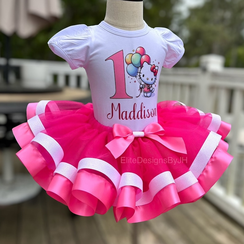 May include: A pink tulle tutu skirt with white satin ribbon trim. The skirt is decorated with a pink bow. A white t-shirt with a Hello Kitty graphic and the number 1 is worn over the skirt. The shirt is personalized with the name "Maddison".