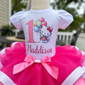 May include: A white t-shirt with a pink number one and a Hello Kitty graphic holding balloons. The shirt has the name Maddison printed below the graphic. The shirt is paired with a pink tulle tutu with a pink satin bow.